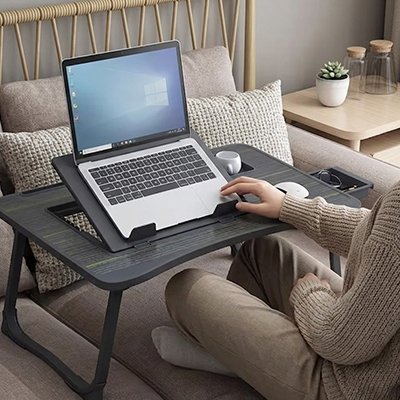 Personne utilisant la table ordinateur portable ComfortStation⹠depuis son canapé avec un ordinateur portable posé sur le plateau inclinable. La table ergonomique comprend une surface ventilée, un support pour smartphone, un espace de rangement intégré et une conception pliable. Solution idéale pour le télétravail, les études, la lecture ou le divertissement à domicile tout en conservant une posture confortable.