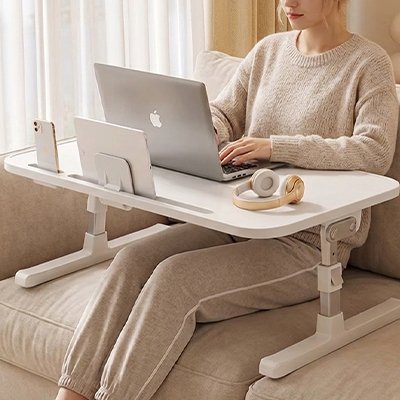 Personne utilisant une table pour ordinateur portable réglable installée sur un canapé. Le plateau ergonomique accueille un ordinateur portable, une tablette, un smartphone et des accessoires audio. Grâce à sa hauteur ajustable et son design moderne, cette table de lit et de canapé offre un espace de travail confortable pour le télétravail, les études, la lecture ou le divertissement à domicile.