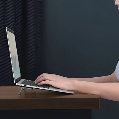 Support bureau pour ordinateur portable pliable en aluminium utilisé avec un ordinateur portable sur un bureau en bois. Son design compact et ergonomique surélève l'écran pour favoriser une meilleure posture, réduire les douleurs au cou et au dos et améliorer la ventilation de l'appareil. Léger, discret et facile à transporter, il est idéal pour le télétravail, le bureau, les études et les déplacements.