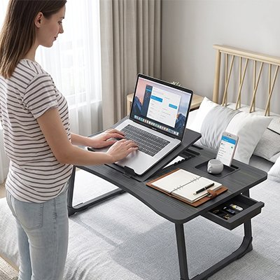 Personne utilisant la table ordinateur portable ComfortStation™ en position debout sur un lit. Le plateau ergonomique accueille un ordinateur portable, un carnet de notes et un smartphone grâce à ses espaces de rangement intégrés. Son support inclinable ventilé améliore le confort de travail, favorise une meilleure posture et transforme facilement un lit en espace de télétravail ou d'étude pratique.