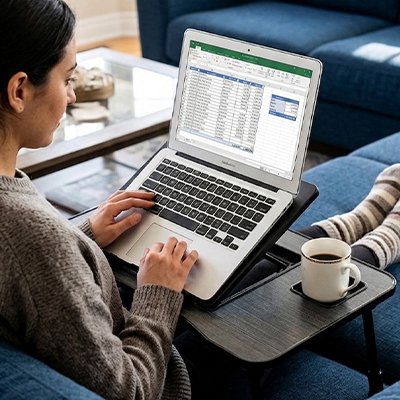 Personne utilisant la table ordinateur portable ComfortStation™ depuis son canapé avec un ordinateur portable posé sur le plateau inclinable. La table ergonomique comprend une surface ventilée, un support pour smartphone, un espace de rangement intégré et une conception pliable. Solution idéale pour le télétravail, les études, la lecture ou le divertissement à domicile tout en conservant une posture confortable.