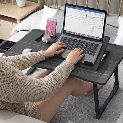 Personne utilisant la table ordinateur portable ComfortStation™ depuis son lit avec un ordinateur portable posé sur le plateau inclinable. La table ergonomique comprend une surface ventilée, un support pour smartphone, un espace de rangement intégré et une conception pliable. Solution idéale pour le télétravail, les études, la lecture ou le divertissement à domicile tout en conservant une posture confortable.